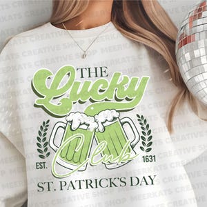 May include: White long-sleeved shirt with green text that reads "The Lucky Club" and "St. Patrick's Day". The shirt also features a green shamrock design and two green beer mugs.