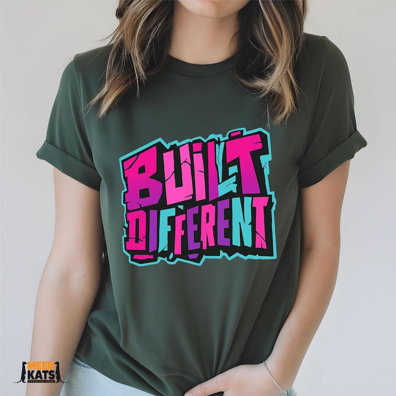 May include: Dark green t-shirt with the words "BUILT DIFFERENT" in a graffiti-style design. The text is in shades of pink, purple, and blue with a black outline. The shirt has rolled-up sleeves.