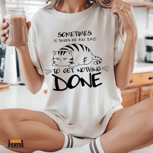 May include: Cream-colored t-shirt with a black and white graphic of a cat and the text "Sometimes it takes me all day to get nothing done." The cat is lying down with a grumpy expression.