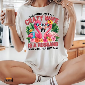 May include: Cream-colored t-shirt featuring a graphic of two pink flamingos wearing sunglasses, surrounded by flowers and tropical leaves. The text reads "BEHIND EVERY CRAZY WIFE IS A HUSBAND WHO MADE HER THAT WAY."