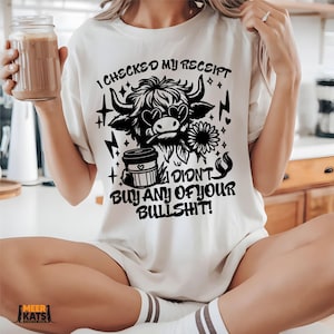 May include: Cream-colored t-shirt with a black graphic of a Highland cow wearing heart-shaped sunglasses, a sunflower, and the text "I CHECKED MY RECEIPT I DIDN'T BUY ANY OF YOUR BULLSHIT!". A coffee cup and lightning bolts are also featured.