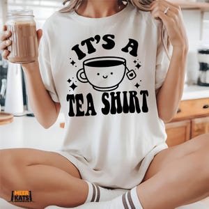 May include: Cream-colored t-shirt with the text "IT'S A TEA SHIRT" in black, featuring a smiling teacup graphic. The shirt is a casual, short-sleeved design, perfect for tea lovers. The shirt is made of a soft material.