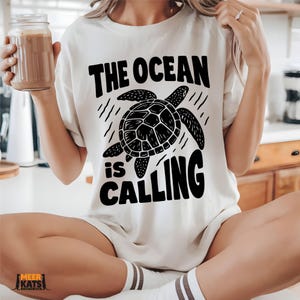 May include: Cream-coloured t-shirt with the text "THE OCEAN IS CALLING" in bold black letters. A black turtle illustration is centered on the shirt. The shirt is being worn.