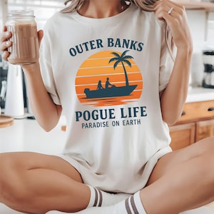 May include: Cream-colored t-shirt with the words "Outer Banks Pogue Life Paradise on Earth" in navy blue. The design features a sunset with a palm tree and a boat with two figures. The shirt is a casual, relaxed fit.