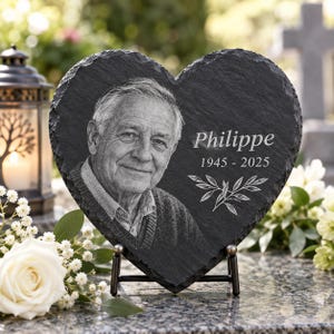 Puede incluir: Una placa conmemorativa de pizarra negra en forma de corazón con el retrato grabado de una persona mayor, el nombre Philippe, las fechas 1945-2025 y un diseño decorativo de rama de hojas. La placa se exhibe en un soporte de metal negro.