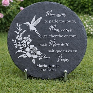 May include: A round, dark gray slate memorial plaque with white text and a hummingbird and floral design. The plaque includes the name "Maria James" and dates "1942-2024". It is displayed on a metal stand.