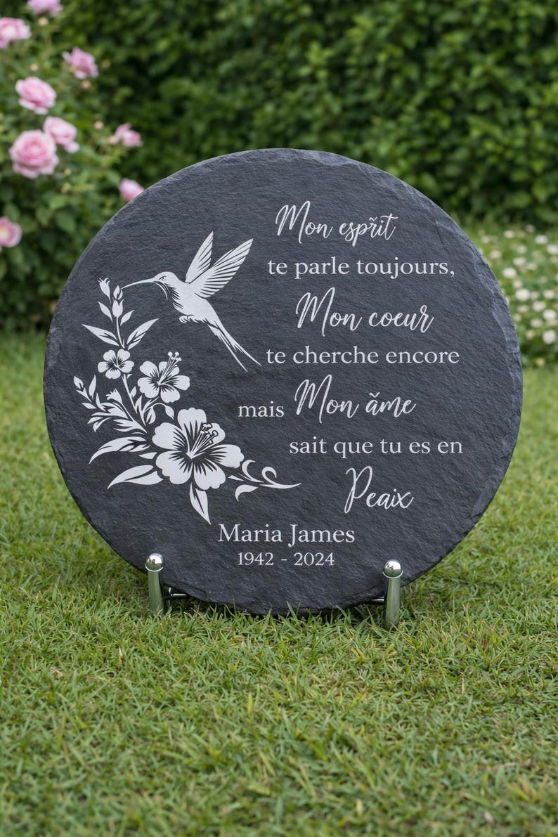 May include: A round, dark gray slate memorial plaque with white text and a hummingbird and floral design. The plaque includes the name "Maria James" and dates "1942-2024". It is displayed on a metal stand.
