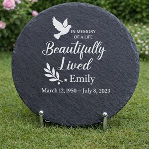 May include: A round, black slate memorial stone with white text and a dove graphic. The text reads "IN MEMORY OF A LIFE Beautifully Lived Emily March 12, 1950 &ndash; July 8, 2023." The stone is displayed on a metal stand.