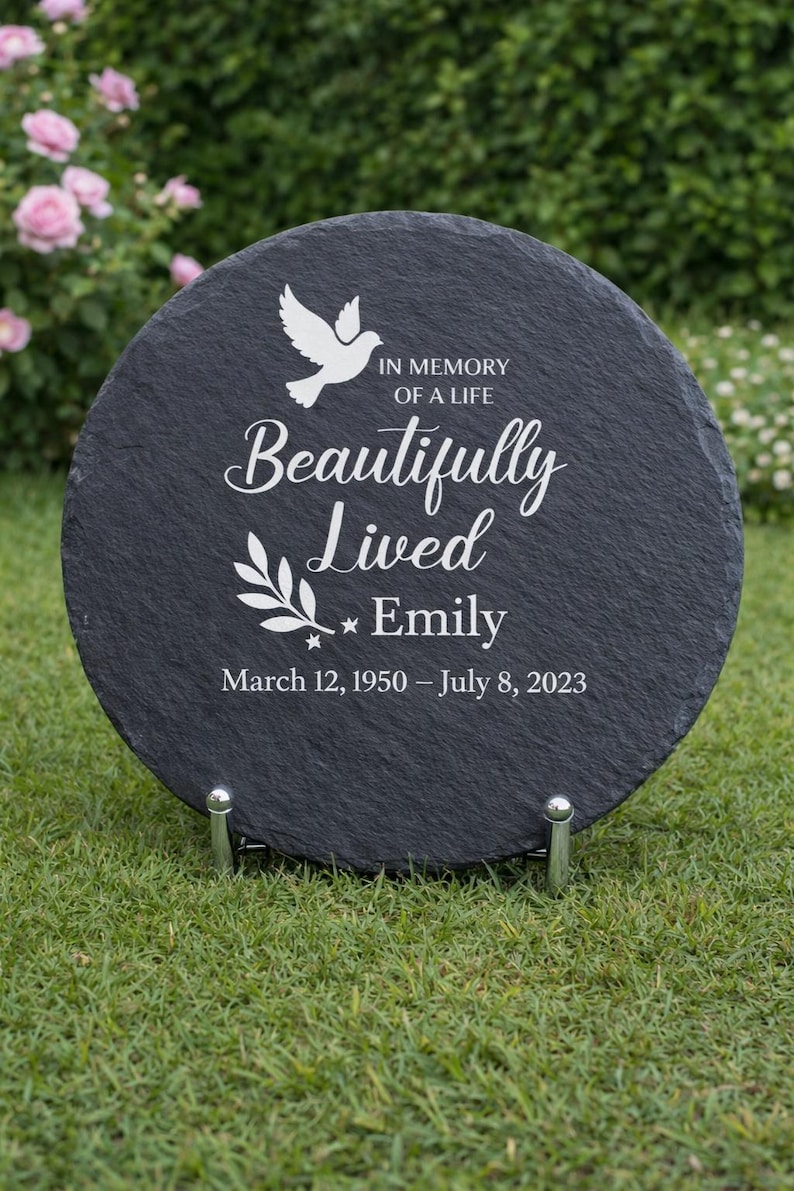 May include: A round, black slate memorial stone with white text and a dove graphic. The text reads "IN MEMORY OF A LIFE Beautifully Lived Emily March 12, 1950 &ndash; July 8, 2023." The stone is displayed on a metal stand.