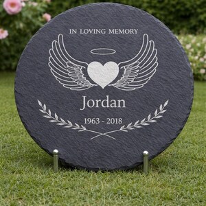 May include: A round, dark gray memorial stone with the inscription "IN LOVING MEMORY." It features a heart with angel wings, the name "Jordan," and the years "1963-2018." The stone is supported by two metal stands and set on a grassy lawn.