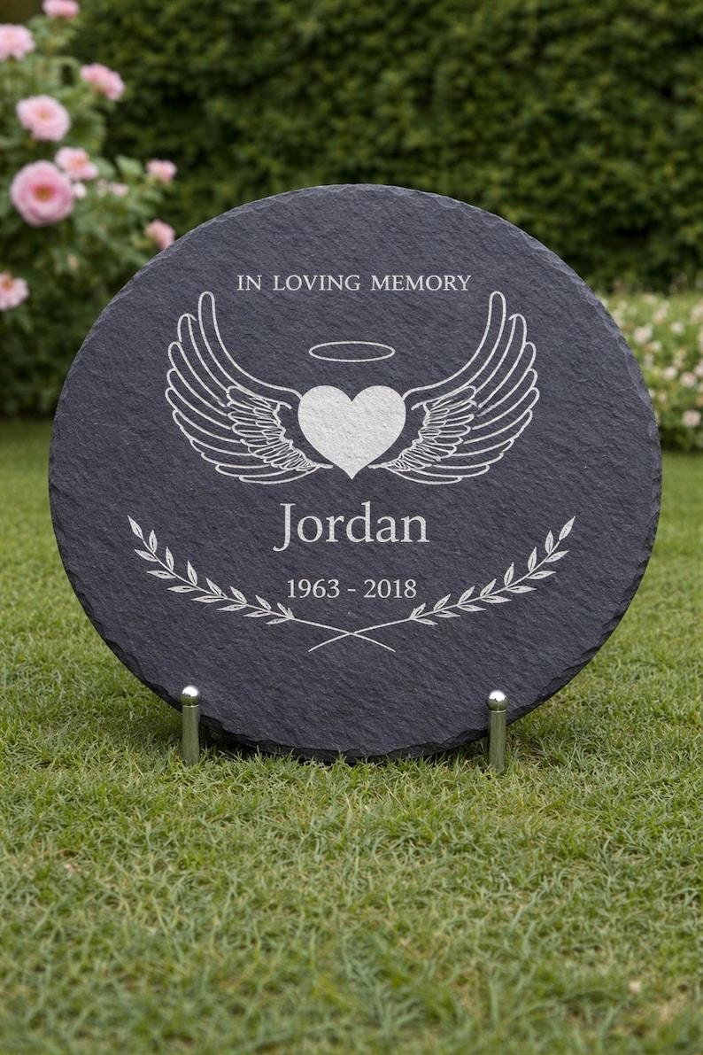 May include: A round, dark gray memorial stone with the inscription "IN LOVING MEMORY." It features a heart with angel wings, the name "Jordan," and the years "1963-2018." The stone is supported by two metal stands and set on a grassy lawn.
