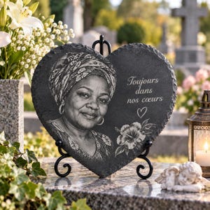 May include: A heart-shaped black slate memorial plaque featuring an engraved portrait of a person wearing a headwrap, the French text "Toujours dans nos cœurs," a small heart symbol, and a hibiscus flower, displayed on a black metal stand.