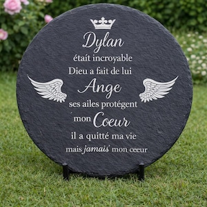 May include: A round, black slate memorial plaque with white text and a crown and wing design. The text reads "Dylan", followed by a French message. The plaque is displayed on a black stand, set against a backdrop of green grass and flowers.