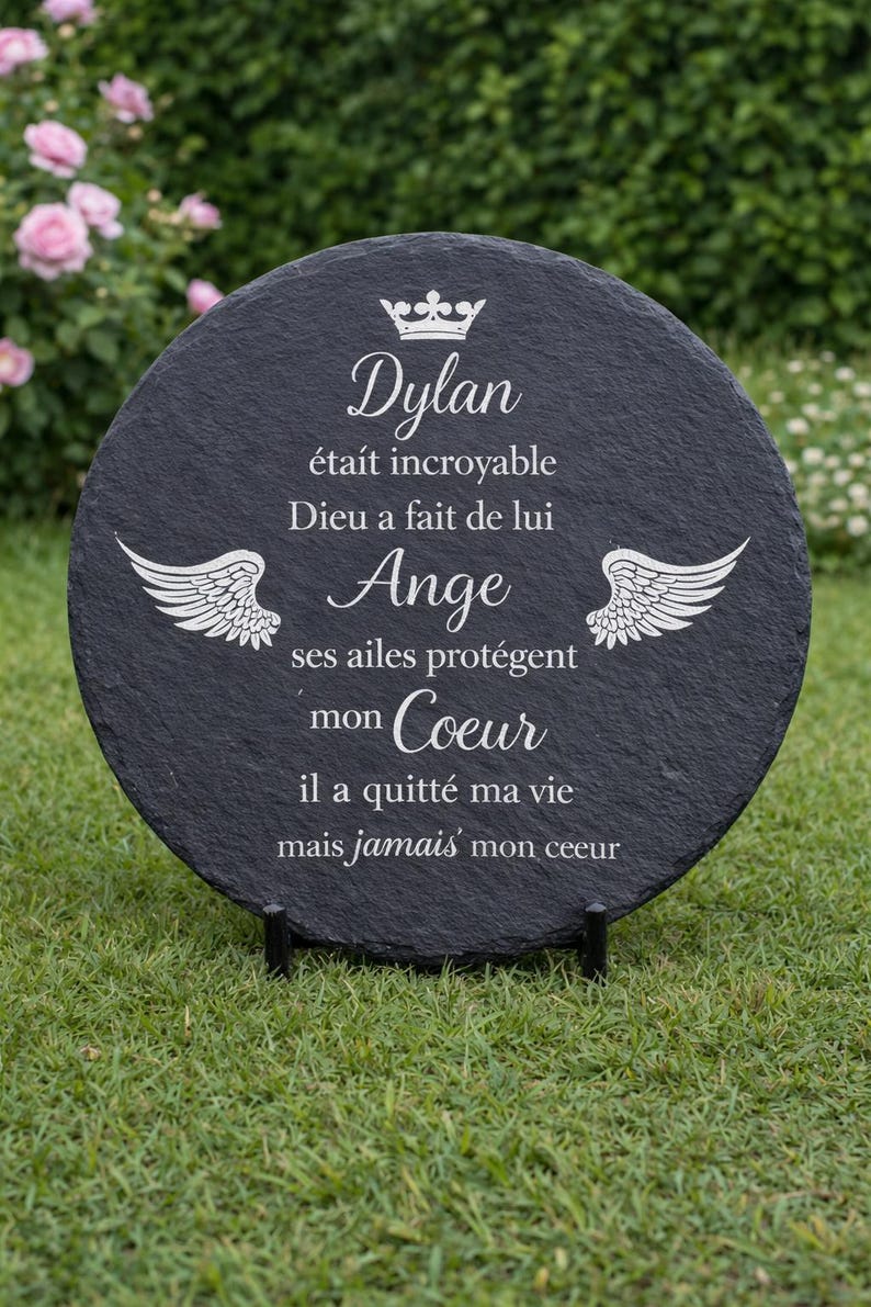 May include: A round, black slate memorial plaque with white text and a crown and wing design. The text reads "Dylan", followed by a French message. The plaque is displayed on a black stand, set against a backdrop of green grass and flowers.