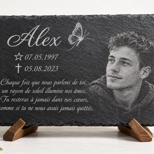 Pode incluir: Uma placa memorial retangular de ardósia preta com um retrato gravado de uma pessoa jovem, o nome Alex, uma borboleta, as datas 07.05.1997 e 05.08.2023 e texto em francês. A placa é apoiada por dois pequenos suportes de madeira.