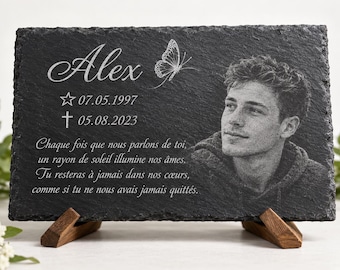 May include: A rectangular black slate memorial plaque featuring an engraved portrait of a young person, the name Alex, a butterfly, dates 07.05.1997 and 05.08.2023, and French text. The plaque is supported by two small wooden stands on a white surface.
