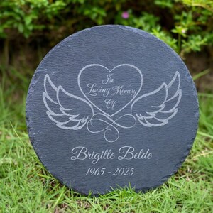 May include: A round, dark gray slate memorial stone with engraved text and a heart-and-wings design. The text reads "In Loving Memory Of" above the name "Brigitte Belde" and the years "1965-2025".