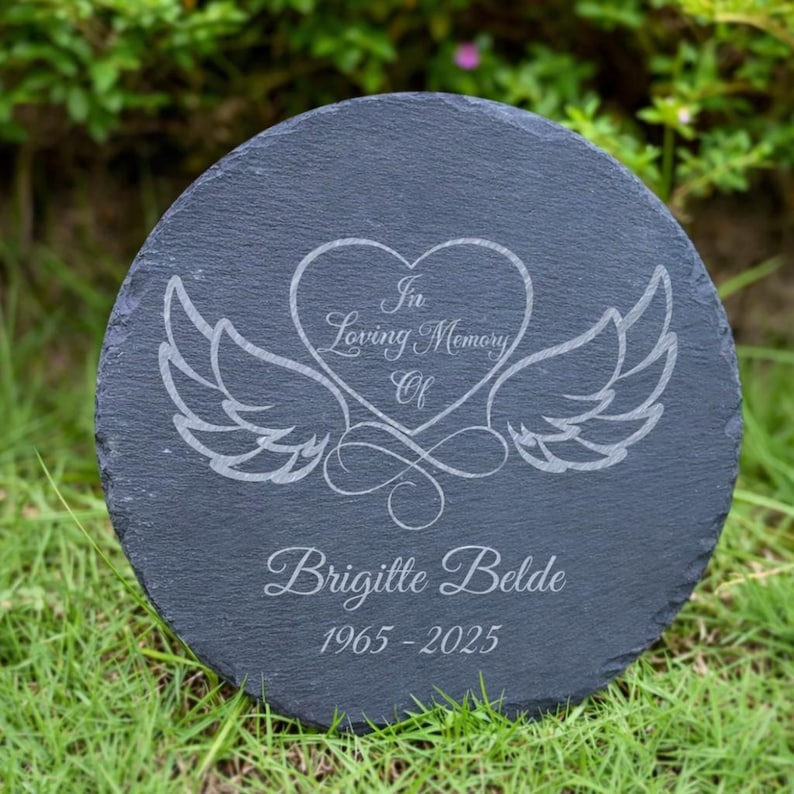 May include: A round, dark gray slate memorial stone with engraved text and a heart-and-wings design. The text reads "In Loving Memory Of" above the name "Brigitte Belde" and the years "1965-2025".