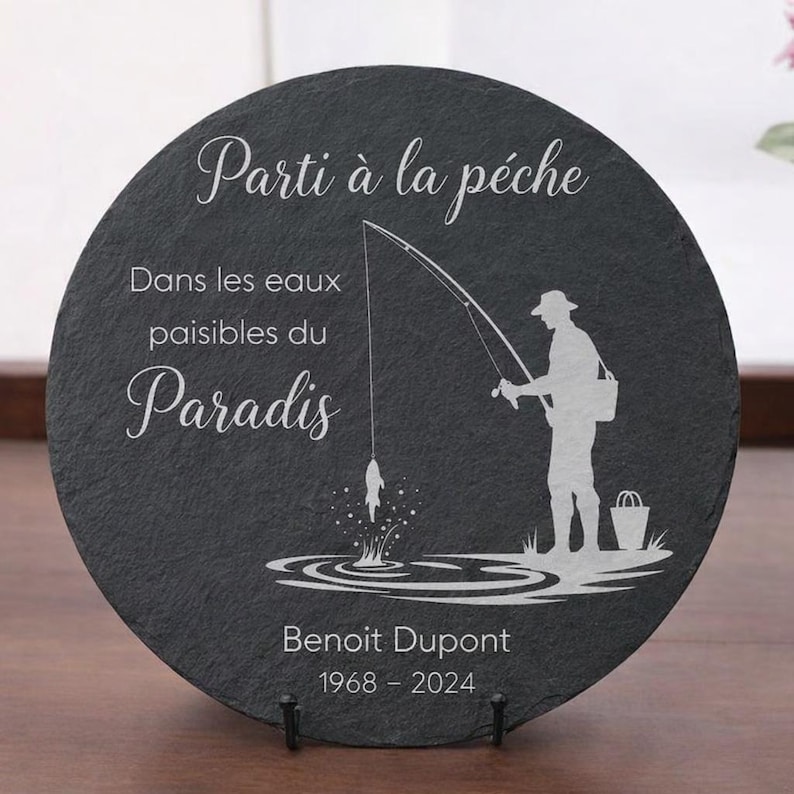 May include: A round, dark gray slate stone with white etched text and a fisherman silhouette. The text reads "Parti &agrave; la p&ecirc;che", "Dans les eaux paisibles du Paradis", and the name "Benoit Dupont" with dates 1968-2024.