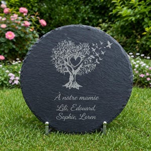 May include: A round, black slate memorial plaque with a tree design, a heart-shaped cutout, and flying birds. The text "&Agrave; notre mamie" is above the names Lili, Edouard, Sophie, and Loren, all engraved in a delicate font. The plaque is displayed on a metal stand.