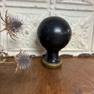 Może przedstawiać: Czarny, kulisty uchwyt do szafki z mosiężną podstawą stoi na drewnianej powierzchni. Suche kwiaty ostu w odcieniach fioletu i brązu znajdują się po lewej stronie uchwytu. Tło przedstawia białą, teksturowaną ścianę.