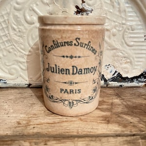 May include: A vintage ceramic jar with a label that reads "Confitures Surfines Julien Damoy PARIS". The jar is decorated with floral designs and has a light brown colour.