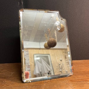 Pode incluir: Um espelho vintage retangular com uma borda chanfrada e uma forma ligeiramente irregular. O espelho tem uma moldura prateada e reflete uma sala com piso de madeira e um guarda-chuva branco.