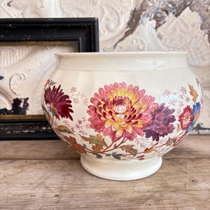 Può includere: Ciotola in ceramica color crema con una forma ampia e arrotondata. La ciotola è decorata con grandi motivi floreali nei toni del rosa, rosso, viola e giallo. I fiori sono ambientati su uno sfondo di foglie e steli verdi. La ciotola poggia su una base bianca.