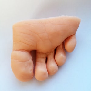 May include: A close-up of a realistic-looking, flesh-toned, silicone foot model with four toes. The foot is slightly angled and the toes are curled.