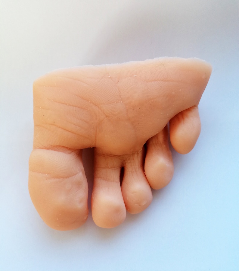 May include: A close-up of a realistic-looking, flesh-toned, silicone foot model with four toes. The foot is slightly angled and the toes are curled.