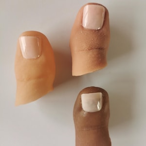 Può includere: Tre coperture protesiche per dita dei piedi in varie tonalità di pelle, ognuna dotata di un'unghia artificiale liscia e chiara. I pezzi in silicone sono sagomati per adattarsi al dito, mostrando texture cutanee realistiche e sfumature.