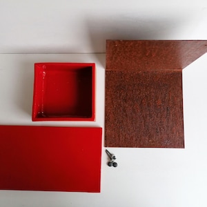 Puede incluir: Una caja de madera roja con tapa y una base roja separada. La caja está hecha de madera y tiene un acabado liso. La caja está sobre una superficie blanca. Hay dos pequeñas piezas de metal negro en la superficie.