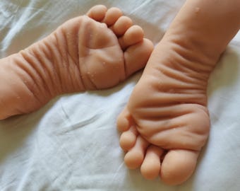 May include: A pair of realistic silicone baby feet with light skin tones, featuring detailed skin texture, visible wrinkles, and soft rounded toes. The feet are positioned side by side, showcasing a lifelike appearance with subtle creases.