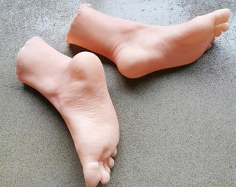 May include: Two flesh-toned silicone foot models on a gray concrete surface. The feet are posed as if walking, with one foot in front of the other.