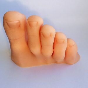 May include: A close-up of a light pink, fleshy, realistic-looking foot with five toes. The toes are slightly curled and the foot is positioned as if it is standing on its side.