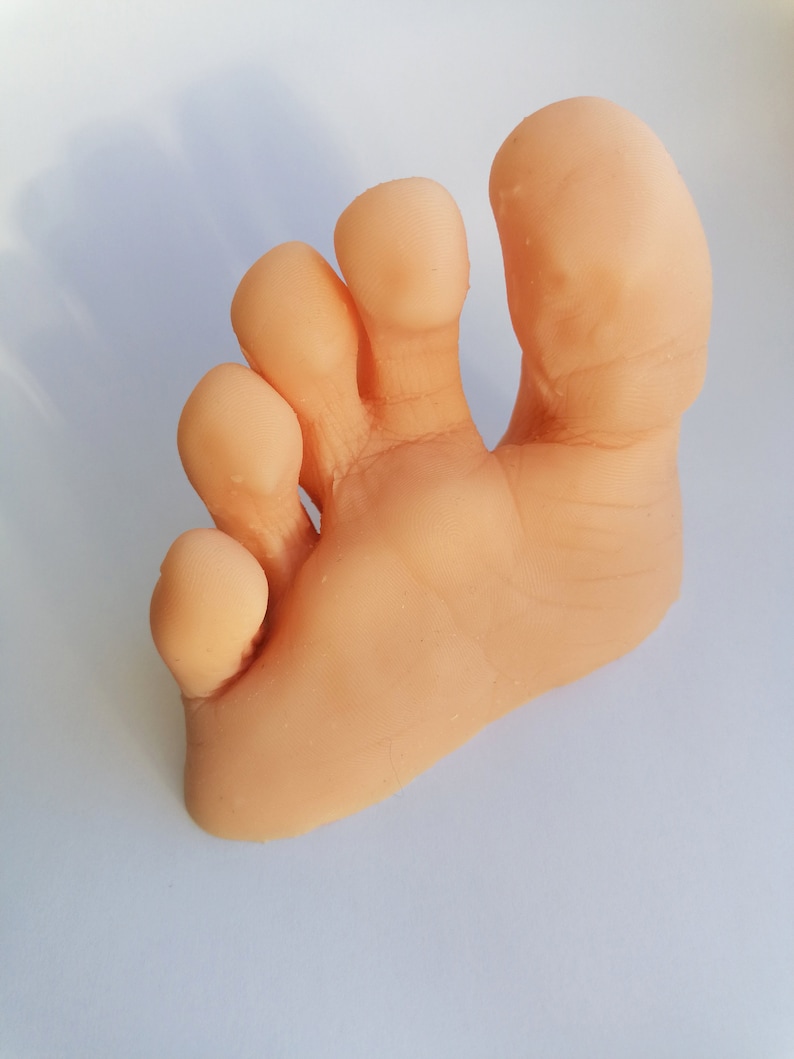May include: A close-up of a light pink, fleshy, human foot with five toes. The toes are curled and the foot is resting on a white surface.