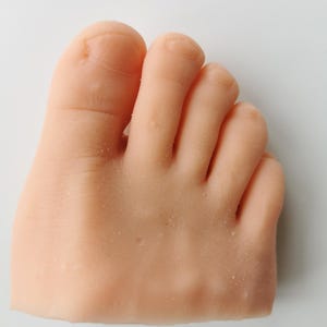 May include: A close-up of a light-colored, realistic-looking foot model. The foot is made of a smooth, soft material and has five toes.