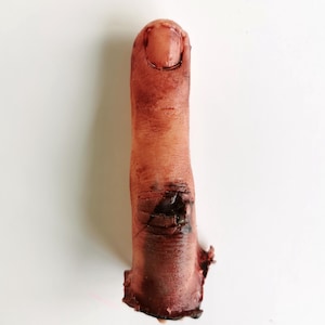 Puede incluir: Un dedo medio protésico en tono carne, con una decoloración oscura, similar a la sangre, cerca de la base. La uña es visible. El texto en la parte superior dice: "Male middle finger. Hollow, wearable." El texto "Variation 1" está en la parte inferior.