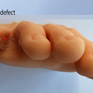 May include: Close-up of a light-colored silicone hand model with a casting defect on the back of the hand. The defect is a small, raised area with a rough texture.