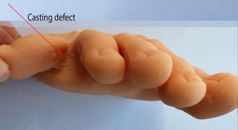 May include: Close-up of a light-colored silicone hand model with a casting defect on the back of the hand. The defect is a small, raised area with a rough texture.