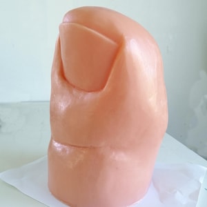 Pode incluir: Um close-up de um dedo do pé humano esculpido, de cor de carne, realista, com uma unha.