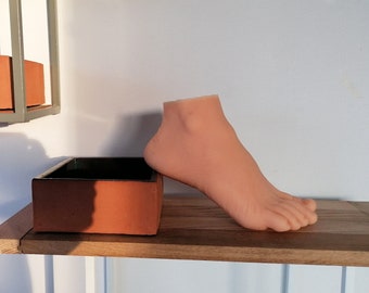 May include: A light brown ceramic planter with a black interior sits on a wooden shelf. A nude-colored foot mannequin is leaning against the planter.