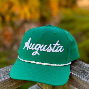 Könnte beinhalten: Eine leuchtend grüne Baseballkappe mit dem in weißer Schreibschrift gestickten Wort Augusta auf der Vorderseite. Die Kappe verfügt über ein weißes, geflochtenes Kordeldetail, das horizontal am unteren Rand der Krone über dem flachen Schirm verläuft.