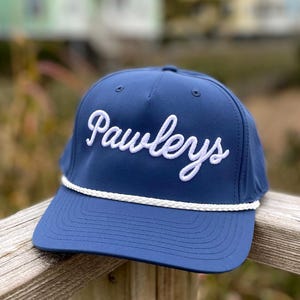以下が含まれることがあります： 白い筆記体で「Pawleys」の文字が刺繍された、ネイビーブルーの野球帽。つばに沿って白いロープのディテールがあり、木製の表面に置かれています。