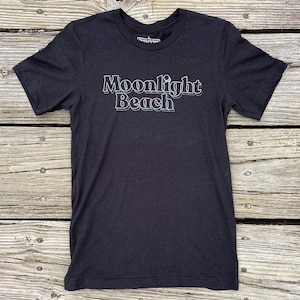 May include: Dark gray t-shirt with the words "Moonlight Beach" in a white and light blue font. The shirt is made of a soft material and has short sleeves. The shirt is displayed on a wooden surface.
