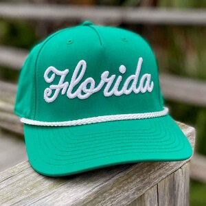 May include: A green baseball cap with the word "Florida" embroidered in white script across the front. The cap has a white rope detail above the brim and a structured fit. The hat is displayed on a wooden surface.