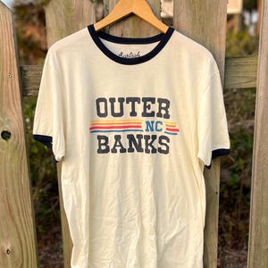May include: A cream-coloured ringer t-shirt with dark navy blue trim on the collar and sleeves. The front features the text "OUTER BANKS" in dark navy blue, with "NC" and horizontal red, yellow, and blue stripes between the words.