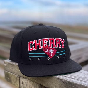 May include: A black baseball cap featuring the embroidered red text CHERRY above a red silhouette of South Carolina with a white crescent moon and palmetto tree. Below the state shape are two horizontal teal lines and the white text GROVE.