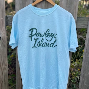 Puede incluir: Camiseta azul claro con el texto "Pawleys Island" en escritura verde oscuro. La camiseta está colgada de una percha de madera contra una valla de madera desgastada. El fondo está borroso, lo que sugiere un entorno al aire libre.