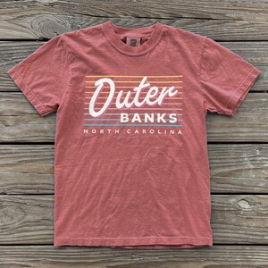 May include: A dusty rose short-sleeve t-shirt featuring a graphic with the text Outer Banks North Carolina in white script and block lettering, layered over a series of horizontal lines in yellow, orange, light blue, and white colors.